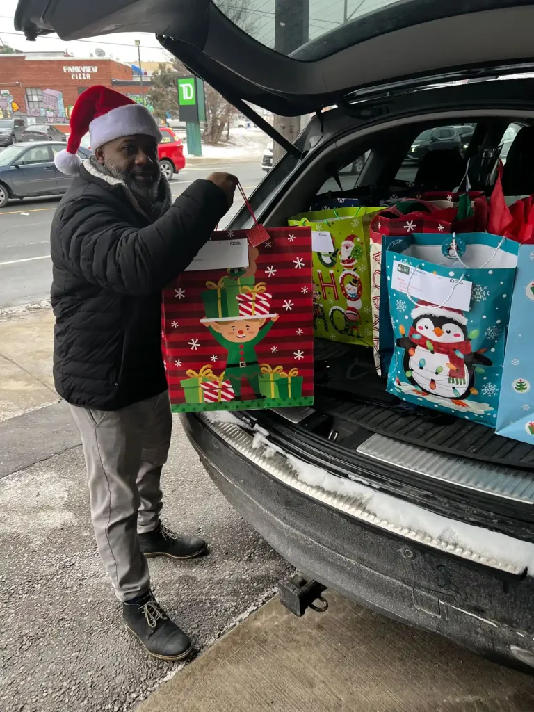 Allan Skeete wearing Santa hat loading colorful holiday gift bags into vehicle trunk on Toronto street for Be A Santa To A Senior deliveries