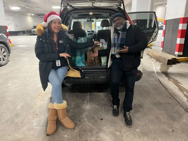 Home Instead Toronto East staff and Trisura representative standing beside vehicle loaded with holiday gift bags in parking garage